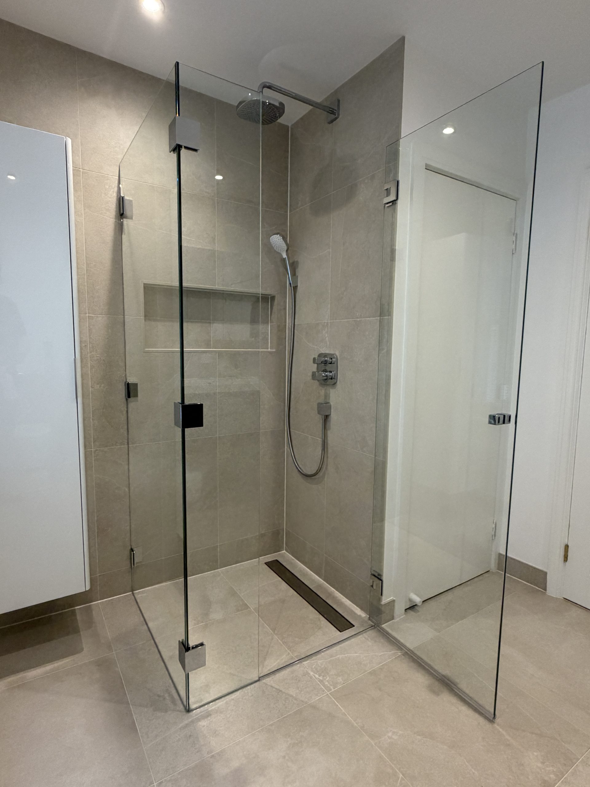 Frameless glass shower door enclosure with a rainfall showerhead and handheld wand in a gray tiled bathroom.