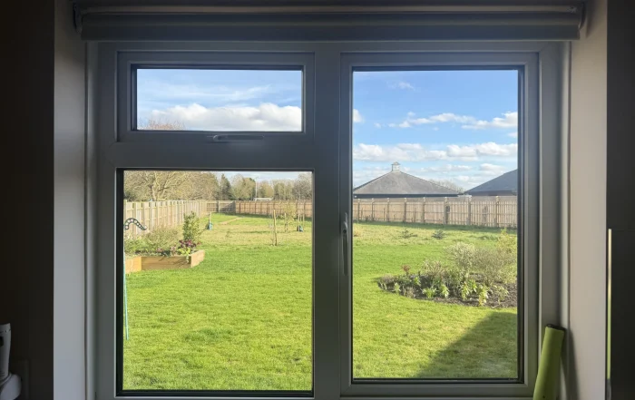 Interior shot of a white-framed window opening to a sunny backyard with a wooden fence, green lawn, and trees under a blue sky with clouds